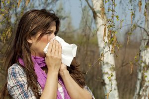A photograph of a woman suffering from allergies blowing her nose, with trees in the background.